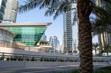 Şık gökdelenler, palmiye ağaçları ve çağdaş mimarinin yer aldığı modern bir şehir manzarası. Sahne, açık mavi gökyüzüne karşı cam ve çelik yapıların bir karışımını yakalıyor..