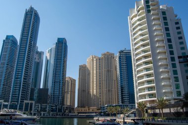 Dubai Marina 'daki modern gökdelenlerin çarpıcı görüntüsü, önplanda açık mavi gökyüzünün altında mimari tarzlar ve lüks yatların karışımını sergiliyor..