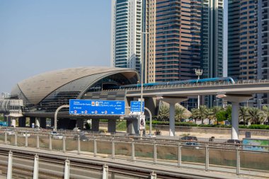 Pürüzsüz, kıvrımlı çatı tasarımlı, yüksek gökdelenlerle çevrili modern bir metro istasyonu. İstasyona bir metro treni yaklaşıyor, birden fazla dilde işaretleri var. Palmiye ağaçları bölgeyi kaplıyor ve şehir manzarasına yeşillik katıyor..