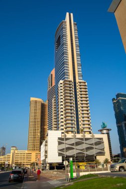 Modern bir gökdelen, diğer binalarla çevrili açık mavi bir gökyüzüne karşı dimdik ayakta duruyor. Mimaride cam ve beton karışımı bir modern tasarım sergilenmektedir. Ön planda birkaç araba ve yayanın bulunduğu bir sokak da yer alıyor.