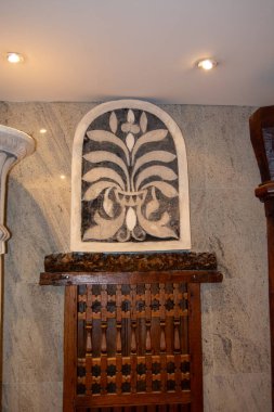 Dekoratif bir duvar özelliği, siyah beyaz çiçeklerle süslenmiş, desenli taştan bir arka planda. Altında, karmaşık oymalar olan ahşap bir panel estetik çekiciliğe katkı sağlar..