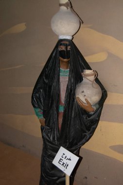 Geleneksel Orta Doğu kıyafeti giymiş bir manken, elinde bir çömlek tutarken diğerini kafasında dengeliyor. Manken, Arapça ve İngilizce metni olan bir çıkış işaretinin yanına yerleştirildi..