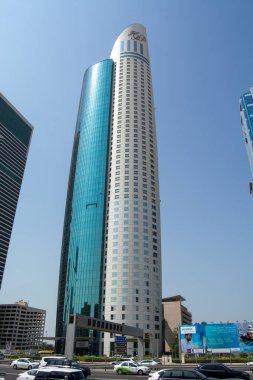 Masmavi gökyüzünü yansıtan parlak cam cepheli, uzun, modern bir gökdelen. Binanın eşsiz kıvrımlı bir şekli var ve işlek bir caddede diğer binalar ve araçlarla çevrili..