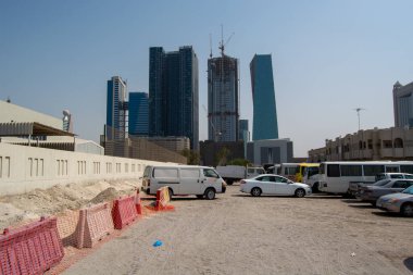 Arka planında modern gökdelenler olan bir kentsel alanda inşaat alanı. Araçlar kumlu bir alana park edilmiş, önlerinde inşaat bariyerleri var..