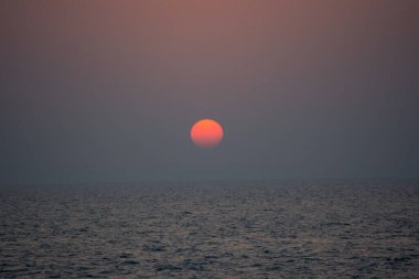 Okyanusun üzerinde sakin bir günbatımı, ufukta kısmen görünen büyük bir güneş, sisli bir gökyüzüne sıcak bir turuncu parıltı yayıyor..