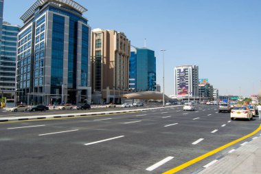 Uzun cam binalar, geniş araçlı bir yol ve açık mavi gökyüzünün yer aldığı modern bir kentsel sokak manzarası. Mimari, ticari alanların bir karışımıyla çağdaş tasarımı sergiler..