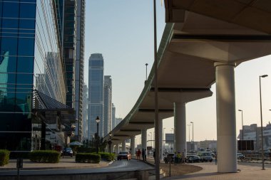 Uzun gökdelenler ve otoyol üstgeçitlerinin olduğu modern bir şehir manzarası. Görüntü cam binalar ve beton yapılar arasındaki zıtlığı yakalıyor, arabalar ve yayalar aşağıda görünüyor.