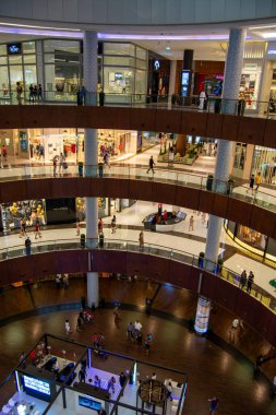 Birçok katlı modern bir alışveriş merkezinin havadan görünüşü. Alışverişçiler her katta görülür, dükkanlar ve vitrinler görünür. Tasarım, ahşap aksanlar ve parlak ışıklandırma içeriyor, canlı bir atmosfer yaratıyor..