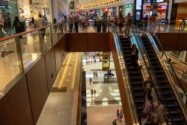 Yürüyen merdivenlerin, alışverişçilerin ve çeşitli perakende mağazalarının olduğu işlek bir alışveriş merkezi. Modern mimari cam parmaklıklar ve parlak ışıklandırmayı içeriyor, canlı bir atmosfer yaratıyor..
