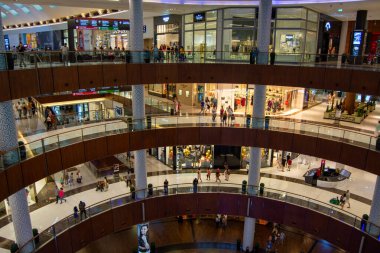 Çok katlı bir alışveriş merkezi. Çeşitli katlarda müşteriler var. Modern bir mimari tasarım sergiliyorlar. Mekan hareketli, dükkanlar ve yürüyen merdivenlerle dolu..
