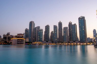 Alacakaranlıkta sakin bir su kütlesine yansıyan modern gökdelenlerin çarpıcı görüntüsü. Ufuk çizgisi, binaları aydınlatan ışıklarla mimari tarzların bir karışımını sergiliyor..