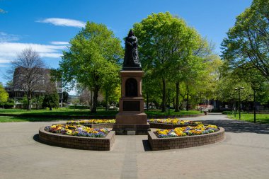 Parkta renkli çiçeklerle çevrili bir heykel. Bereketli yeşil ağaçlar gölge sağlar ve berrak mavi gökyüzü sakin atmosferi güçlendirir. Heykele giden yollar ziyaretçileri keşif yapmaya davet ediyor..