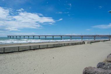 Okyanusa doğru uzanan uzun bir rıhtımın manzarası, kıyıya vuran dalgalar. Gökyüzü parlak mavi, dağınık bulutlar ve kumlu plaj ön planda görülebilir..
