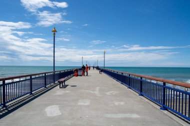Parlak mavi gökyüzünün altında dağınık bulutlarla okyanusa doğru uzanan uzun bir iskelenin manzarası. İnsanlar rıhtımda yürüyor, deniz kenarındaki atmosferin tadını çıkarıyorlar..