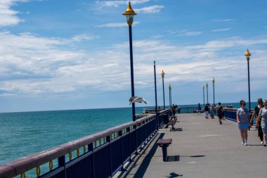 Okyanusa doğru uzanan bir rıhtımın manzarası. Yürüyüş yolunda yürüyen insanlar. Gökyüzü parçalı bulutlu ve bir martı iskelenin kenarında uçuyor..