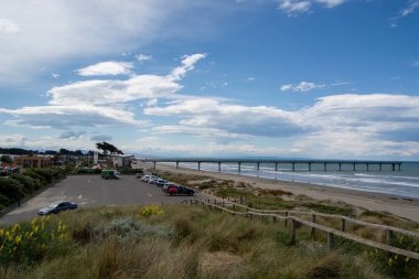 Okyanusa doğru uzanan uzun bir iskelesi olan sahilin manzarası. Ön planda çimenli kum tepeleri ve kır çiçekleriyle çevrili birkaç arabalı bir otopark yer alıyor. Gökyüzü parçalı bulutlu, mavi ve beyaz bulutlar sakin bir atmosfer yaratıyor.
