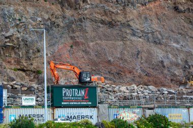 Turuncu bir kazıcı kayalık bir uçurumun yakınındaki bir inşaat sahasına yerleştirildi. Sitede Protranz Dünya Taşımacılığı için robotik yıkım hizmetlerini gösteren büyük yeşil bir tabela bulunuyor. Nakliye konteynırları arka planda görünür..