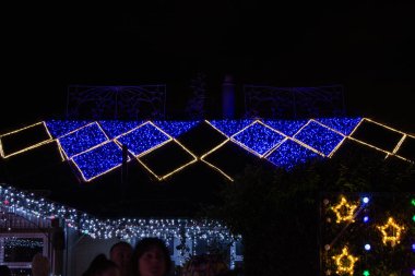 Çatıda mavi ve sarı Noel ışıklarıyla dekore edilmiş bir ev. Geceleri şenlikli bir atmosfer yaratıyor. Arkaplanda ayrıca yanıp sönen ışıklar ve dekorasyonlar yer alıyor..
