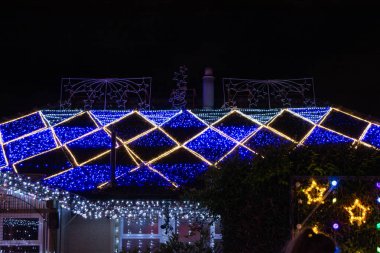 Renkli Noel ışıklarıyla süslenmiş, çatıda mavi ve beyaz elmas desenli, yıldız ve ağaçlar gibi dekoratif elementlerle donatılmış bir ev..