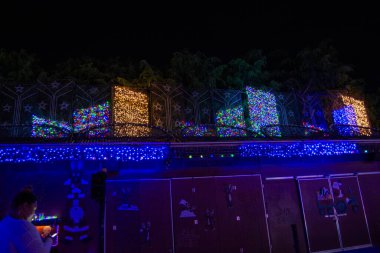 Arkasında karanlık bir gece gökyüzü olan bir çiti süsleyen renkli Noel ışıklarının canlı bir gösterisi. Işıklar neşeli bir atmosfer yaratacak şekilde düzenlenmiş..