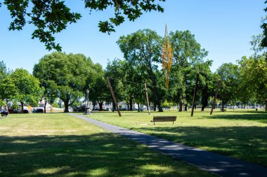 Çimlerin ve ağaçların arasından geçen bir patikanın yer aldığı güneşli bir park sahnesi. Yol boyunca ahşap bir bank var. Arka planda benzersiz bir heykel var..