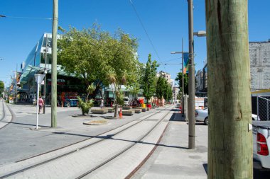 Tramvay rayları, ağaçlar ve modern binaların yer aldığı güneşli bir şehir manzarası. Bölge hareketlilikle dolup taşıyor, yeşillik ve altyapı karışımı sergiliyor.