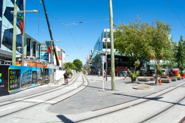 Tramvay raylarının, modern binaların ve ağaçların yer aldığı güneşli bir şehir manzarası. İnsanlar yaya dostu bir bölgede yürürken ve bisiklet sürerken görülüyor. İnşaat çalışmaları arka planda görünür.
