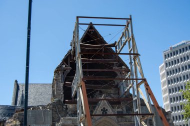 Önünde bina iskelesi olan kısmen çökmüş bir kilise binası hasar görmüş çatı ve yapıyı gözler önüne seriyor. Arka planda modern binalar ve açık mavi bir gökyüzü yer alıyor..