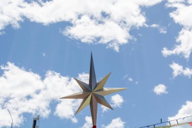 Pofuduk beyaz bulutlarla dolu parlak mavi gökyüzüne karşı keskin noktaları olan büyük, dekoratif bir yıldız. Yıldız belirgin bir şekilde gösterilir ve neşeli bir atmosfer yaratır..