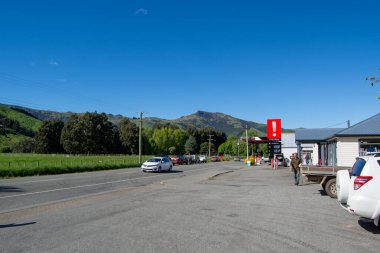Sağında yeşil tepeler ve mavi gökyüzüyle çevrili bir benzin istasyonu olan kırsal bir yolun manzarası. Arabalar yol boyunca park halinde, ve bir kişi benzin istasyonunun yanında duruyor..