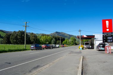 Kenara park etmiş arabaların bulunduğu kırsal bir yol, bir benzin istasyonuna çıkıyor. Açık mavi gökyüzünün altında yemyeşil tepeler var. Benzin istasyonunda belirgin bir tabela var.