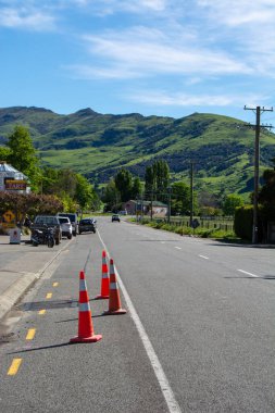 Açık mavi gökyüzünün altındaki yeşil tepelere doğru uzanan turuncu trafik konileriyle kaplı sessiz bir kırsal yol. Manzara huzurlu bir kırsal atmosferi yakalıyor..