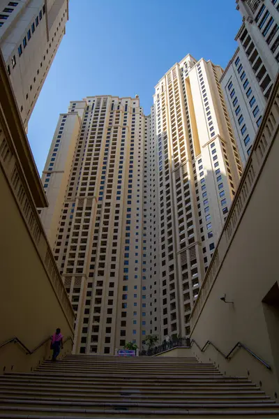 Bir merdivenin tabanından yukarıdaki gökdelenlere bakan bir manzara. Binalar modern ve üzerinde açık mavi gökyüzü olan bej bir cephe içeriyor..