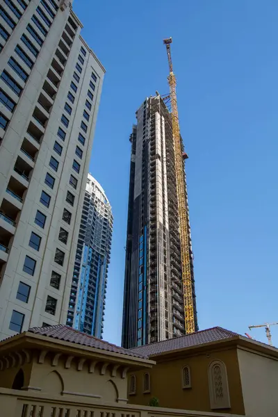 Modern binalarla çevrili, yapım aşamasındaki uzun bir gökdelenin yer aldığı bir inşaat alanı manzarası. Gökyüzü açık ve mavi, gökdelenin tepesinde bir vinç görünüyor..