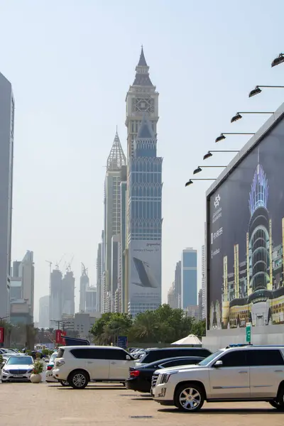 Önemli bir saat kulesi de dahil olmak üzere yüksek binaların yer aldığı modern bir şehir silueti manzarası. Ön planda birçok araba park halinde ve büyük bir reklam görülüyor. Hava açık ve güneşli..