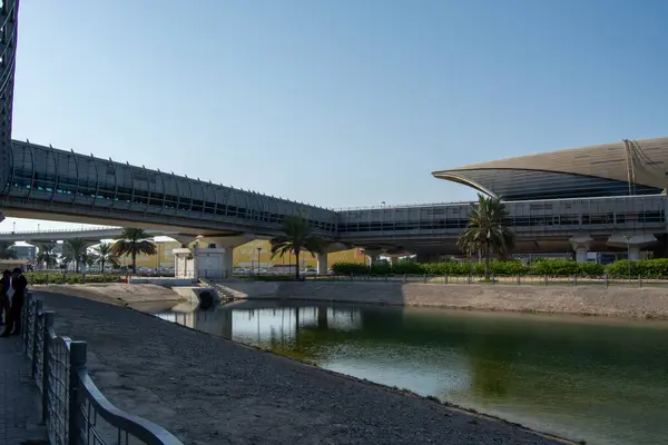 Yüksek yürüme yolları ve sakin bir su kütlesinin yanında zarif bir bina tasarımının yer aldığı modern bir mimari bakış açısı. Palmiye ağaçları bölgeyi kaplıyor, sakin bir atmosfer yaratıyor..