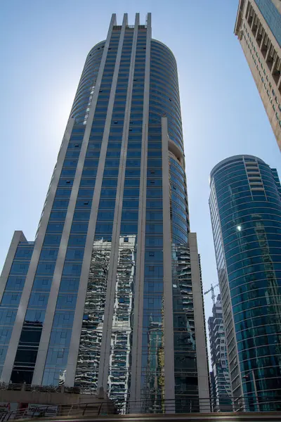 Diğer binalarla çevrili, yansıtıcı cam cepheli, uzun, modern bir gökdelen. Güneş arka planda parlıyor, çarpıcı bir siluet etkisi yaratıyor..