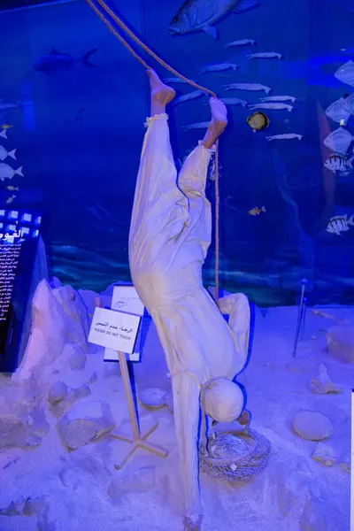 Gerçek boyutta bir dalgıç heykeli baş aşağı asılı, geleneksel dalış kıyafetleri giyiyor, yanında sepet var. Arka planda balık ve deniz yaşamının yer aldığı bir sualtı sahnesi yer alıyor..