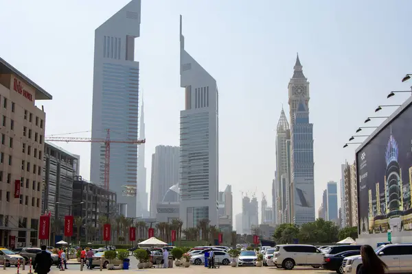 Modern bir şehirde, eşsiz mimari tasarımları olan gökdelenlerin sergilendiği hareketli bir şehir manzarası. Saat kulesi de dahil. Ön planda arabalar ve yayalar yer alırken, palmiye ağaçları caddeyi kaplıyor.