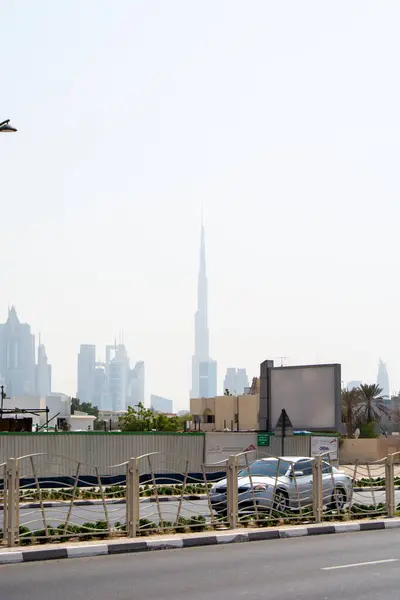 Dubai 'deki Burj Halife' nin uzak bir manzarası, modern gökdelenler ve puslu bir gökyüzü ile çevrili. Ön planda, şehir manzaralarında tipik bir şekilde arabası ve çiti olan bir yol yer alıyor..