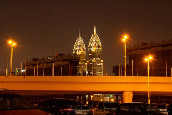 İki uzun, aydınlık gökdelenin gece manzarası. Sivri uçlu, şehir manzarası ile çevrili. Ön planda park halindeki arabaları ve sokak lambaları olan bir otoyol yer alıyor..
