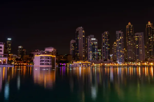 Sakin bir su kütlesine yansıyan muhteşem bir gökyüzü manzarası. Aydınlatılmış pencereli uzun gökdelenler canlı bir atmosfer yaratırken, su renkli ışıkları yansıtıyor..