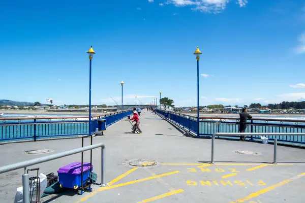 Suya uzanan bir balıkçı iskelesinin manzarası. İnsanların balık tutması ve açık havanın tadını çıkarması. Gökyüzü açık ve mavi, canlı bir atmosfer yaratıyor..