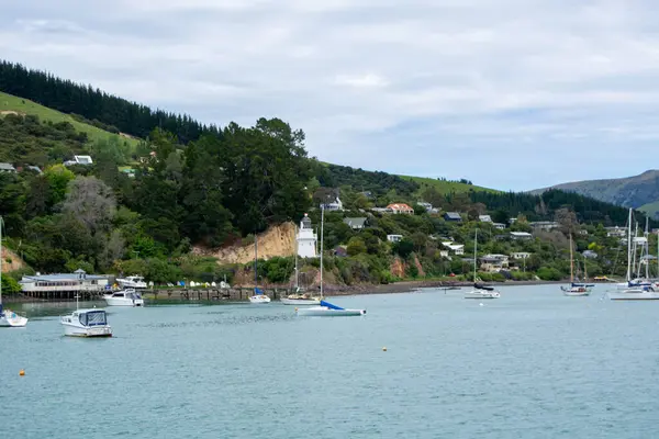 Huzurlu bir sahil sahnesi. İçinde birkaç teknenin bulunduğu sakin bir körfez. Arka planda yemyeşil tepeler yükseliyor, evlerle benekli. Sahilde ağaçlarla çevrili bir deniz feneri duruyor..
