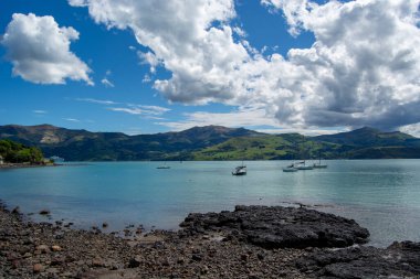Sakin suların, körfezde demirlemiş teknelerin ve arkaplanda parçalı bulutlu bir gökyüzünün altında yemyeşil tepelerin bulunduğu sakin bir kıyı manzarası..
