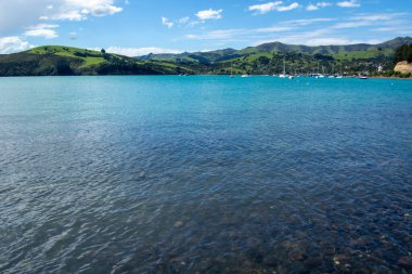 Bereketli yeşil tepeler ve berrak mavi gökyüzü ile çevrili, yumuşak dalgalı turkuaz bir körfez manzarası. Küçük tekneler uzakta demirleyerek, huzurlu bir sahil manzarası yaratıyorlar..