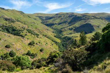 Açık mavi gökyüzünün altında yuvarlanan yeşil tepelerin ve vadilerin manzarası. Manzara bereketli bitki örtüsü, ağaçlar ve dolambaçlı bir vadi içeriyor, doğanın güzelliğini gözler önüne seriyor..