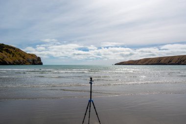 Sahilde hafif dalgaların dalgalandığı sakin bir sahil sahnesi, ön planda bir tripodu var. Gökyüzü parçalı bulutlu ve tepeler arka plandaki kıyı şeridini çerçeveliyor..