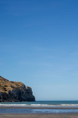 Yumuşak dalgaların sahile vurduğu sakin bir sahil sahnesi. Arka planda kayalık bir uçurum, berrak mavi bir gökyüzünün altında..