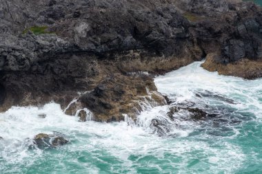 Kıyıya vuran dalgaların oluşturduğu kayalık bir sahil şeridi beyaz köpük oluşturur. Su canlı bir turkuazdır, karanlık kayalarla çelişir. Sahne doğanın gücünü yakalar..